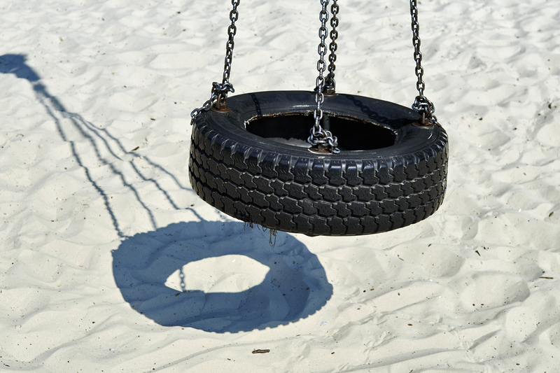 A black rubber tyre repurposed as a swing seat, suspended by metal chains attached to its inner edge, hanging from an unseen support. The tyre has a textured surface with visible tread patterns and a slightly glossy finish. It is positioned slightly above a sandy surface, likely part of a playground or outdoor area, with its shadow cast clearly on the light-colored sand below, creating a circular shape. The sand appears fine and soft with some small pebbles or debris scattered around. The background shows more of the sandy area, with subtle footprints and irregularities in the surface. The overall scene is illuminated by natural daylight, giving a bright and neutral atmosphere. This image is suitable for illustrating garden or outdoor rubbish removal services, especially those that include clearing or maintenance of outdoor play areas or gardens in London or nearby locations, with a focus on environmental responsibility and outdoor cleaning.