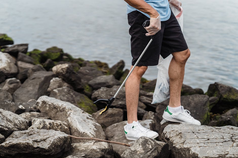 A man standing on a rocky shoreline near a body of water, engaging in a rubbish collection activity. He is wearing a light blue T-shirt, black shorts, and white sneakers with green accents, while his 