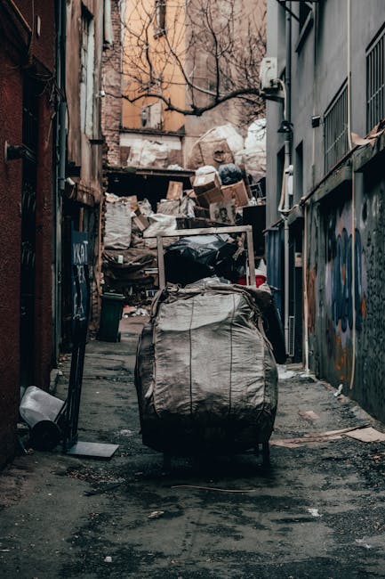 The image depicts the exterior of a building with a textured grey concrete wall and a metal door painted in light grey, situated in an urban environment. Ten blue rubbish bags made of glossy plastic m