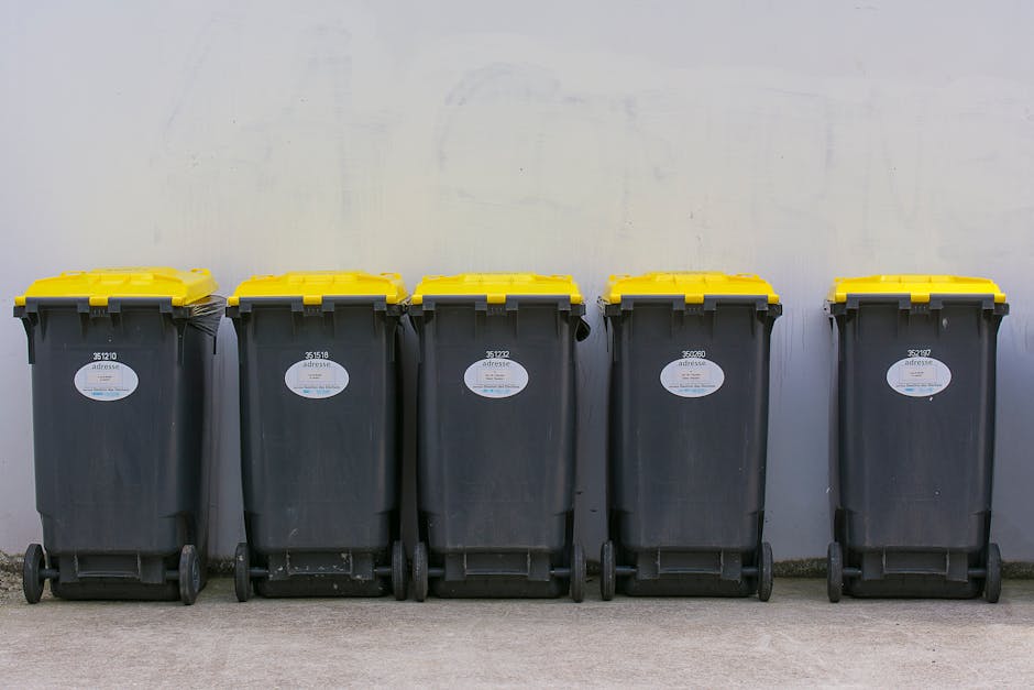 A street scene with a bright green waste collection vehicle facing away from the camera, positioned in the middle of a wide pedestrian pathway. The vehicle’s rear opened to reveal compartments for r