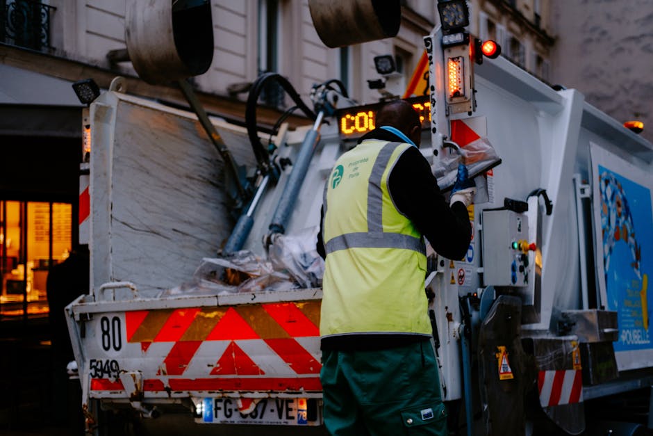 A waste collection worker wearing a high-visibility yellow vest with reflective strips and dark clothing is operating a large, white rubbish truck. The truck is equipped with mechanical lifting and lo